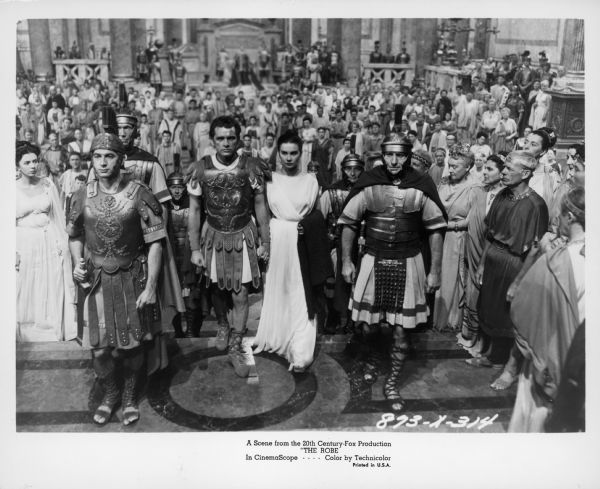Scene still from the film "The Robe." Marcellus Gallio (Richard Burton) and Diana (Jean Simmons) are walking hand in hand in the center. They are surrounded by Roman centurions, and a large crowd of people are in the background.