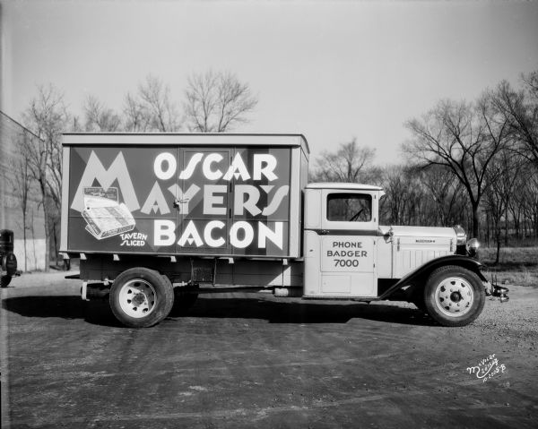 Profile view of the right side of an Oscar Mayer bacon truck made by Diamond T parked near a building. The sign on the back of the truck reads: "Oscar Mayer's Bacon" and "Tavern Sliced."