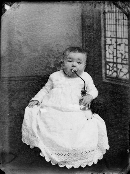 Studio portrait of a baby suckling on a bottle. The child is sitting upright dressed in a white gown, with his right hand resting on the chair arm, and his left hand over a glass bottle with a cork and long, flexible tube with a nipple on the other end which the baby is sucking. The baby has short hair and is facing forward. The painted backdrop depicts a window on the baby's left.
