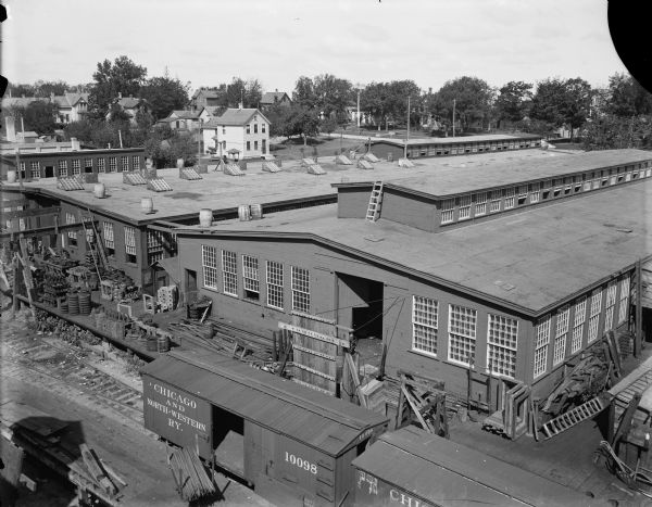 Elevated view of the Fuller & Johnson Mfg. Co., Ltd. The office was at 101-133 North Dickinson St. and these buildings were/are in the 1400 block between East Mifflin Street and East Dayton Street. They became part of the Trachte Company in later years. In the foreground are railroad cars near a loading dock. Painted sign on the side of the railroad car reads: "Chicago and North-Western RY." The two-story house in the upper left is located at 214 North Baldwin Street.