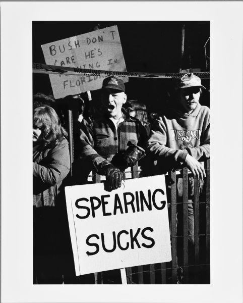 Protesters standing behind a fence at an anti-spearfishing rally. The man in the center is holding a sign that reads: "Spearing Sucks."