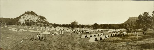 Elevated view of tents at Camp Douglas, with rock formations in the background. Man standing in foreground with hands in his back pockets is Edwin L. Lowndes.