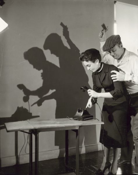 Publicity still for the TV show "Dial 999." A woman is posing dialing a telephone, while a man standing behind her is posing while grabbing her shoulder from behind. The man is preparing to hit her over the head with a gun he is holding in his raised right hand.