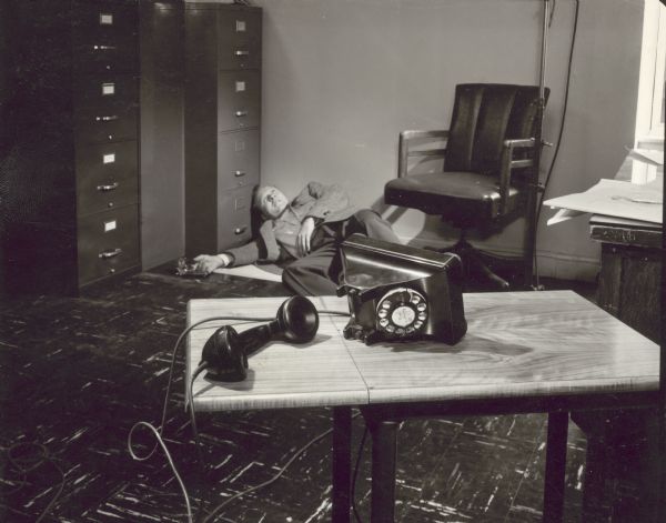 Publicity still for the TV show "Dial 999." A man is lying on the floor against the wall and filing cabinets. There is a gun lying in his right hand. A telephone is tipped over on a table in the foreground.