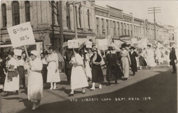 View down street towards women marching in a parade, some carrying signs and some carrying American flags. A number of women are wearing nurse's uniforms. Signs include "6th Ward 100%," "WOMAN'S COMMITTEE Council of "Defense," and "Liberty Loan Committee." Caption reads: "4th Liberty Loan Sept. 28th.