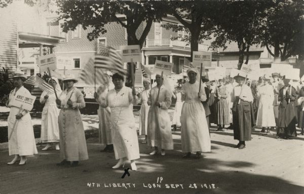 View towards a group of women marching up a street in a parade. Some are wearing nurse's uniforms, some are waving flags, and many are holding signs that read: "WOMAN'S COMMITTEE Council of Defense." Caption reads: "4th Liberty Loan, Sept. 28."