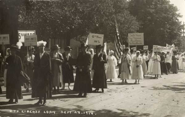A group of women are marching up a street in a parade. Some are carrying signs, and one woman is carrying an American flag. The signs include: "WOMAN'S COMMITTEE Council of Defense," "5TH WARD," "6th Ward 100%," and "Sixth Ward Your Service May Bring The Boys Back." Caption reads: "4th Liberty Loan, Sept. 28."