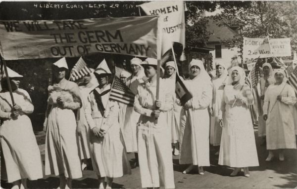 A group of women wearing white nurses' uniforms are marching up a street in a parade. Some are carrying American flags, others are carrying signs and banners that read: "We will Take the Germ out of Germany," "Women of Industry," "Doing Our All — 2nd Ward," and "Third Ward Service." Caption reads: "4 Liberty Loan, Sept 29."