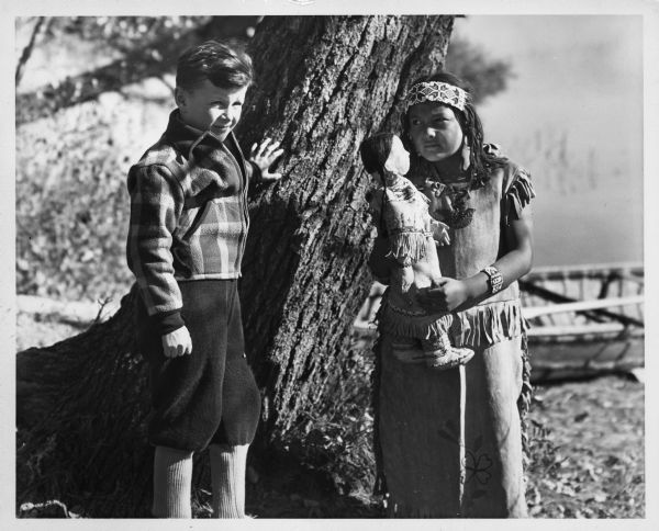 Walker Reid (1929-2016) standing alongside tree with Indian girl. Reid Walker shown is 7-years-old, and is the son of Carol McMillan Reid. Portrait taken at Lac Du Flambeau Reservation. 