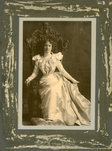 Studio portrait of Mildred wearing a white silk satin dress with corded bodice, while seated in an ornate wooden chair. Her right foot rests on an upholstered foot stool.
