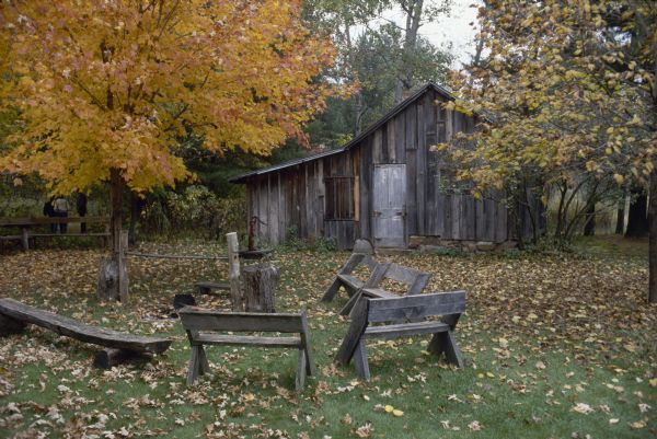 View of Aldo Leopold's shack in the fall. Several Leopold benches are placed around a fire pit in the yard in front of the shack. The is a hand pump near the cabin. People are walking on a path beyond a fence and trees on the left.