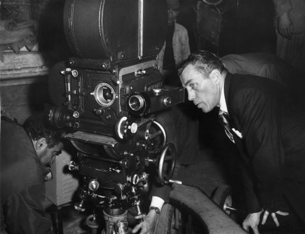 Ed Sullivan is bending down and looking into the eyepiece of a Mitchell movie camera. There are men in the background.