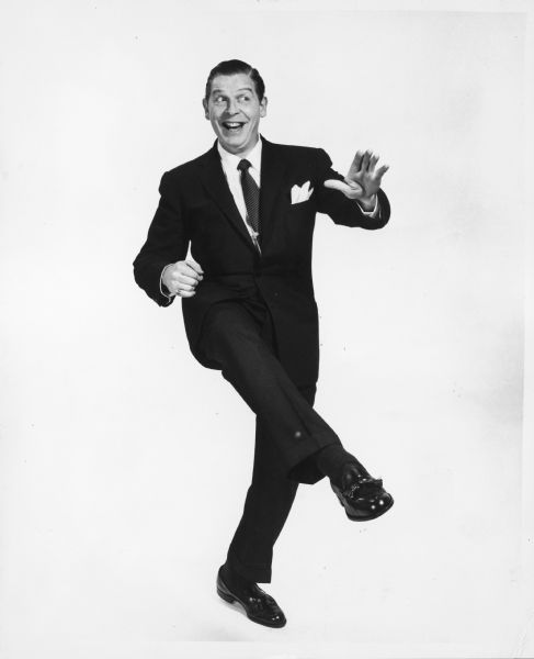 Publicity photograph of Milton Berle posing with one foot in the air and a big smile on his face. He is wearing a dark suit with a white handkerchief in the breast pocket.