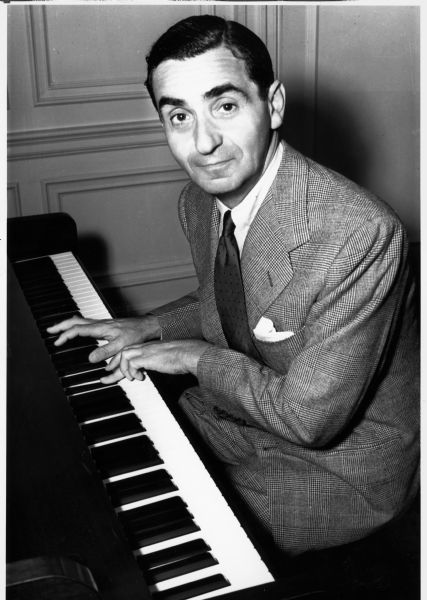 Portrait of Irving Berlin sitting at a piano and looking directly at the camera. He is wearing a suit and tie.