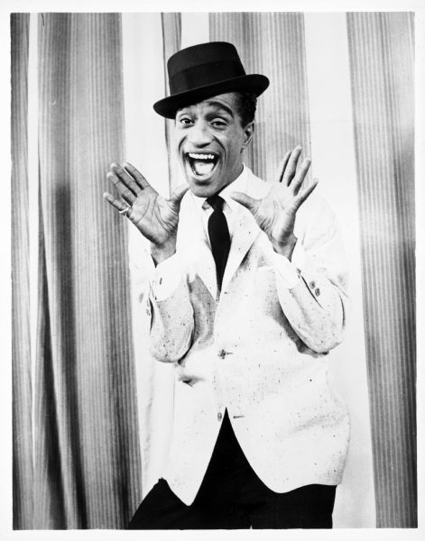 Sammy Davis Jr. posing with his hands up and his mouth open in front of a curtain. He is wearing a suit coat, tie, hat and a pinky ring on one hand.