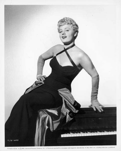 Shelley Winters sitting on top of a piano. She is wearing a long gown and long gloves, and a bracelet. She is smiling seductively at the camera.