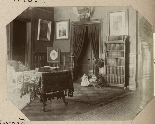 Interior view of the parlor at Oakwood, including a table covered with a tasseled cloth, dolls on a chair, and bookshelves.