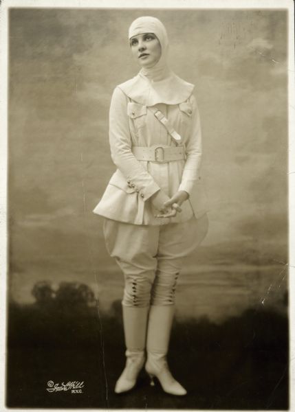 Publicity portrait for the musical revue "Over the Top" of Justine Johnstone dressed as an aviatrix, from head to toe, in an all white outfit with her hands folded in front of her in front of a painted backdrop.
