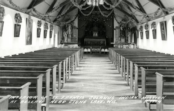 Postcard of the church interior, with pews, altar, nave, statues, and paintings. The church ceiling is decorated with garlands and wreaths. Caption reads: "Interior of St. Francis Solanus Indian Mission at Reserve Near Stone Lake, Wis."