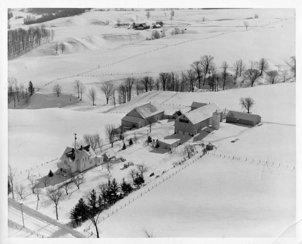 Aerial view of a farm in winter, with the farmhouse, barn, and outbuildings blanketed in snow. The name Cedar Spring Farm is on the barn. Another farm is in the distance. Image caption reads: "Kewaunee (vicinity), Wis. 1955 (?). Photo by George M. Frisbie, De Pere, Wis."