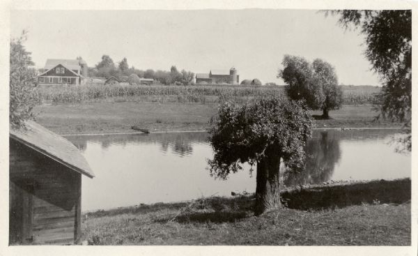 Photographic postcard view of a river, with part of an outbuilding in the foreground on the left. On the opposite shoreline is a farm, with farmhouse, barn, and crop fields. Caption on back reads: "The Bank of the Crawfish, Aztalan, Sept. 1/19."