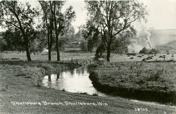 View across field towards a bend in the Shullsburg Branch, which runs from the Galena River near the town of Lead Mine to the town of Shullsburg. A farm is in the background, and a machine in the field on the right, perhaps a thresher, is emitting smoke. Caption reads: "Shullsburg Branch, Shullsburg, Wis."