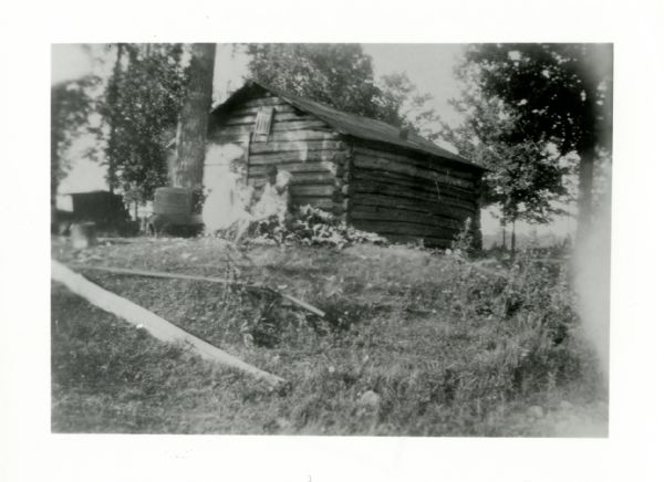 View up hill towards two adults and two children outside of a log building, surrounded by trees. Caption reads: "Sheldon, Wis. 1930s. Log sauna on Matt Johnson farm."