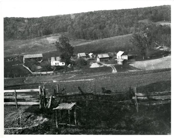 View down hill towards a farm in a valley, with wooded bluffs in the background. In the foreground, a fence is at the top of the hill overlooking the farm. Image caption reads: "Prairie du Sac, Wis. About 1897 (?). Farm. Photo by Chas. N. Brown."