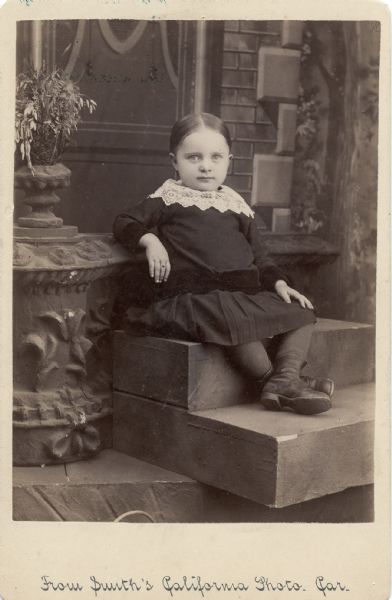 Cabinet card showing a young girl posing on wooden steps on a set. The card is labeled Cora Young on the back. This is possibly Cora Louise Bartlett Young, who was born in Eau Claire in 1900. Ms. Young graduated from the Eau Claire Teachers College, and also studied at UW-Madison and Northwestern. She studied public school music education and served jobs in many towns before settling in Pasadena, California, in the 1930s. There, she served as a music teacher and later as president of the Pasadena Symphony Association, from 1953 to 1961. She was active in many community organizations, including the Red Cross during World War II.