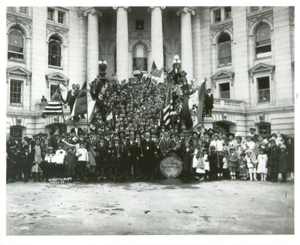 A large group of children and adults are standing outside the Wisconsin State Capitol building, many holding American flags. Some members in the foreground are holding instruments, and a drum reads: "Italian Band. Victorio Emanuel III. Madison - Wis." Caption on the back of the image reads: "Italian "Fourth of July" celebration - 1919[?]." A banner visible in the center-right portion of the image has been identified as belonging to the "Societa di M.S. Bersaglieri." 