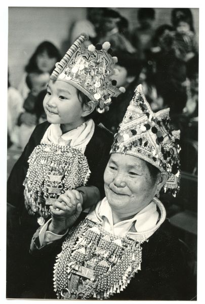 An older woman and a young girl are holding hands and smiling. They are both wearing traditional Hmong jewelry, including the xauv (necklace) and a silver headdress. Caption reads: "GRAND PAIR — Hmong native Zer Yang and her granddaughter Xay Her, watched other performers in the Cultural Exchange Program open house at the Guadalupe Center, 239 W. Washington St. The event, which included ethnic dancing and singing, was part of Wisconsin Electric Power Co.'s energy efficiency demonstration project."