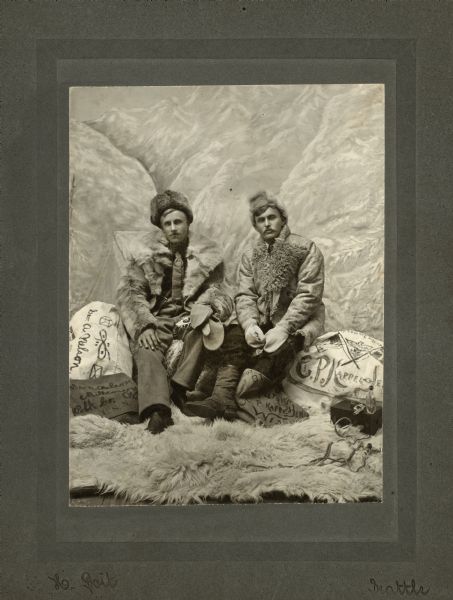 Studio portrait of George A. Nelson (right), and C.P. Kappel dressed in fur hats, coats, and boots. There is a gun and a bottle on top of a small box next to Kappel. This image commemorates their trip to The Yukon to mine gold.