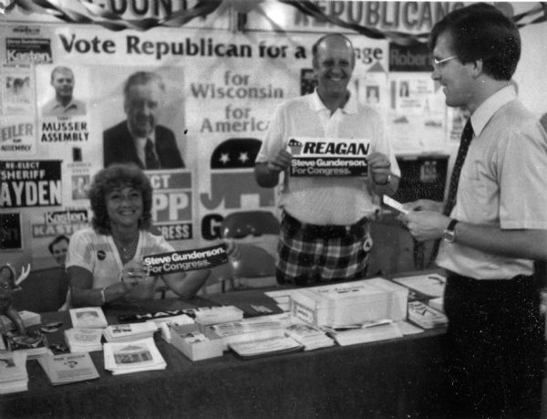 Steve Gunderson at a booth with Republican supporters of his 1980 campaign for U.S. Congress. Campaign materials for several republican politicians are on the table and on the wall in the background. A woman is holding a Steve Gunderson for Congress bumper sticker and a man is holding both a Reagan and a Gunderson bumper sticker.