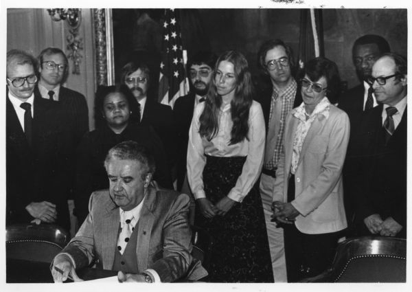 Governor Lee Dreyfus signing a trailer to AB874 (1978), or the Children's Code, as backers of the law look on. Behind Dreyfus left to right: Richard Flintrop, Joel Ungrodt, Marcia Coggs, Rick Phelps, Peter Plau, Eileen Hirsch, unknown woman, unknown man, and Roland Todd.