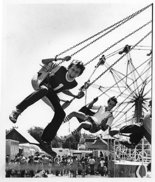 A boy enjoying an amusement ride on swings at the East Side Businessmen's Club carnival at Milwaukee Street and Fair Oaks Avenue. Other riders, carnival-goers and a Ferris wheel are in the background.