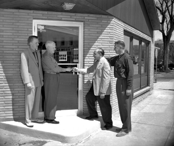 Four men are standing at the door of the Chamber of Commerce on the corner of Oak and Broadway. One of the men is handing a set of keys to another man.