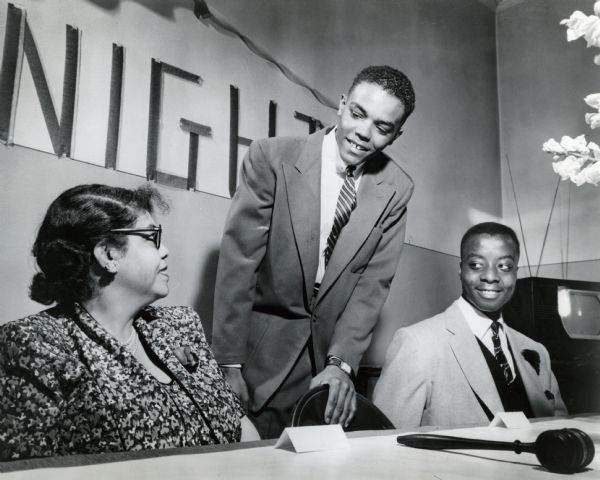 A young man is standing and smiling and looking at a woman sitting on the left, while another young man sitting on the right is looking on. In front of the woman is a gavel, and in the background on the wall is a banner. Caption reads: "Mothers were honored Wednesday night by the Knights Boys Club of Lapham Park Social Center, 1758 N. 9th Street. Mrs. John Givens, 1310 W. Ring Street, heard her son, John, Jr., president of the Knights, give a speech in tribute to mothers. At right is James Reed, 1729 N. 16th Street, vice-president of the Knights."