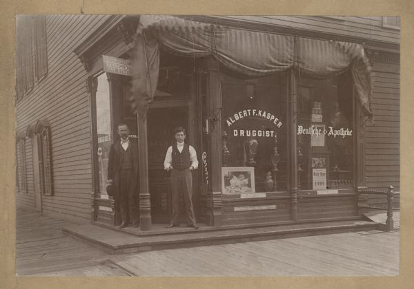 View of two men standing on the steps in front of the entrance of a store on a corner. The sign painted on the window reads: "Albert F. Kasper Druggist," and "Deutsche Apotheke" (German Apothecary). A sign above the door reads: "Public Telephone Pay Station."