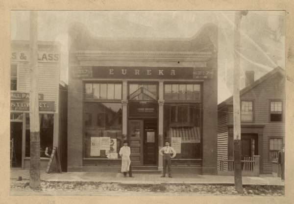 View across street towards two men standing on the board sidewalk in front of the entrance to the Eureka Billiard Hall (217 State Street). Posters in the windows advertise a concert and Volksfest.