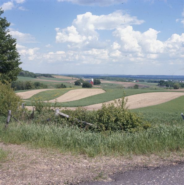 View across fence line towards hilly fields at a dairy farm. A red barn and white silo are in the distance.