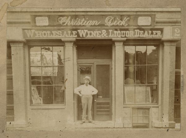 View of Christian W. Dick standing with arms on hips at the entrance to his business on 223 State Street. A poster in the window is advertising Buffalo Bill's Wild West Show.