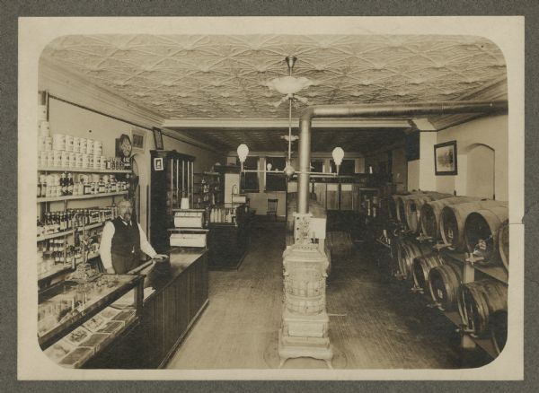 Slightly elevated view of Christian W. Dick, on the left, standing behind the counter of his wine and liquor store. Cigars are in a display case in the left foreground. Kegs of wine and beer are stacked in rows on the right, and assorted goods (such as coffee, condiments, and canned vegetables) are displayed on shelves. A wood burning stove is in the center.