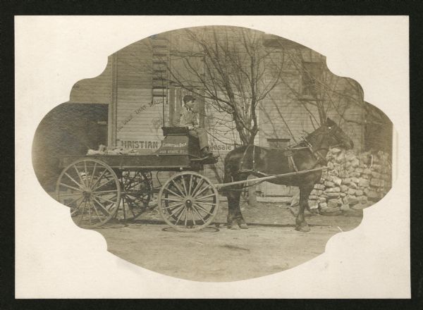 A young man is sitting at the front of a wagon holds the reins of a horse hitched to the cart. Both the cart and building in the background are have painted signs advertising Christian Dick Wholesale Wine and Liquor Store at 223 State Street. The cart is carrying a load of bricks or stone.