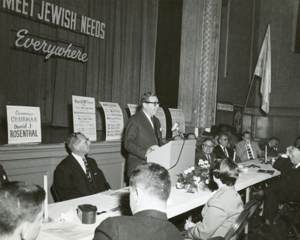 View towards Leo Lania speaking from a podium at the United Jewish Appeal. Men are sitting at the long table on either side of Leo Lania, and men and women are also sitting in the foreground. A sign on the stage reads: "Meet Jewish Needs Everywhere."