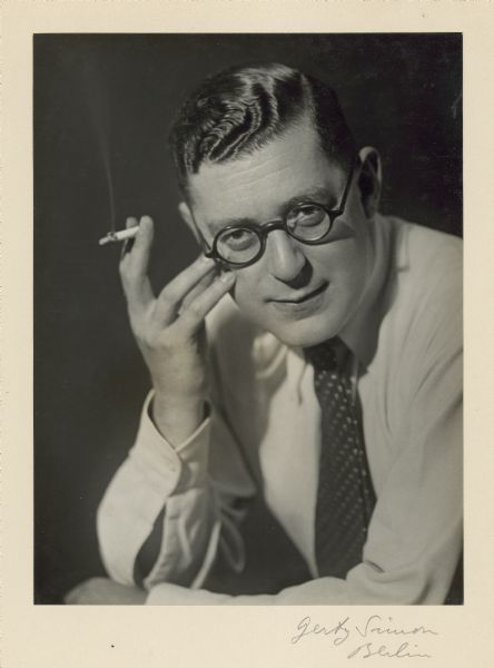 A casual studio portrait of Leo Lania. He is wearing black, round eyeglasses and is smoking a cigarette.