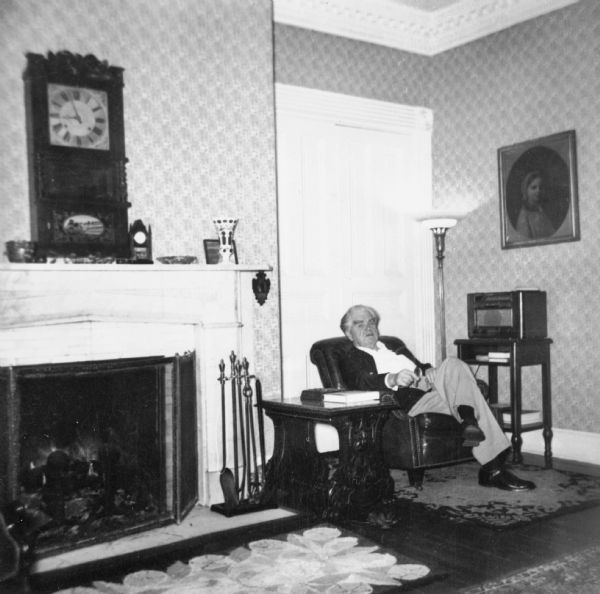 John L. Lewis sitting in his living room next to a fireplace with a clock on the mantle. There is a radio on a stand on the right.