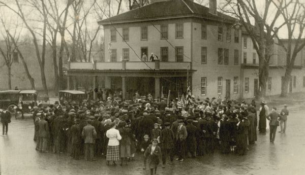 Elevated view of a crowd of people gathering outside a building, some of them carrying American flags. Two men are standing on the balcony, and may be addressing the crowd. Caption reads: "Kilbourn celebrating prematurely over the armistice, Nov. 7, 1918."