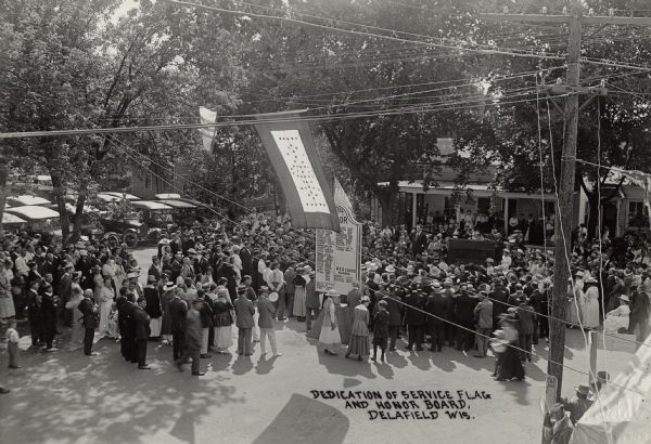 Slightly elevated view of a crowd in a public space. A man wearing a suit is standing on what may be a wagon, and he appears to be addressing the crowd. A flag and an honor roll plaque set up in the middle of the crowd. People are sitting on the porch of a building in the background.