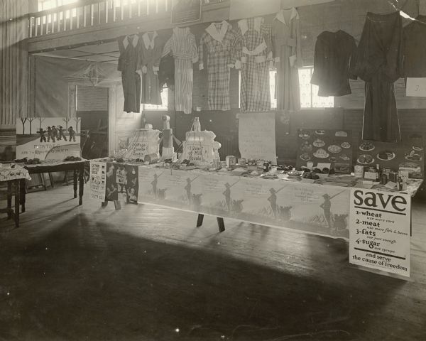 An exhibit inside a building displaying examples of how to make meals for "a patriot's table," as well as the relative value of different foods. A handmade poster reads: "Potatoes are the Home Guard." Several dresses are hanging from the balcony above the table. Caption reads: "Exhibit prepared by the Home Economics Department of the County Council of Defense, Kenosha, Wis. May, 1918."