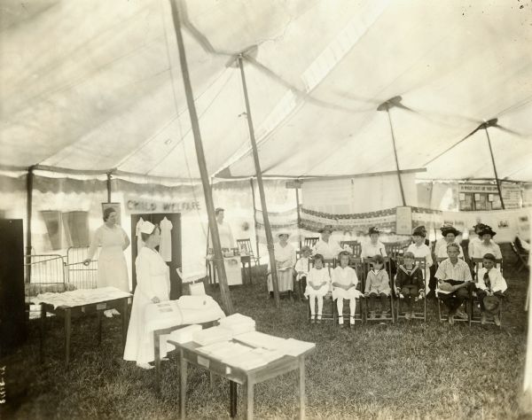 Three women in nurse's uniforms are standing in a tent, where several children and women are seated on chairs. Displays of cribs, cloths (possibly diapers), and baby food are on tables. A sign reads: "Child Welfare." Caption reads: "BABY CLINIC AND REST TENT. DANE CO. FAIR. AUGUST, 1918. Woman's Com[mittee] Dane Co[unty] Council of Defense. Child Welfare Com[mittee]."